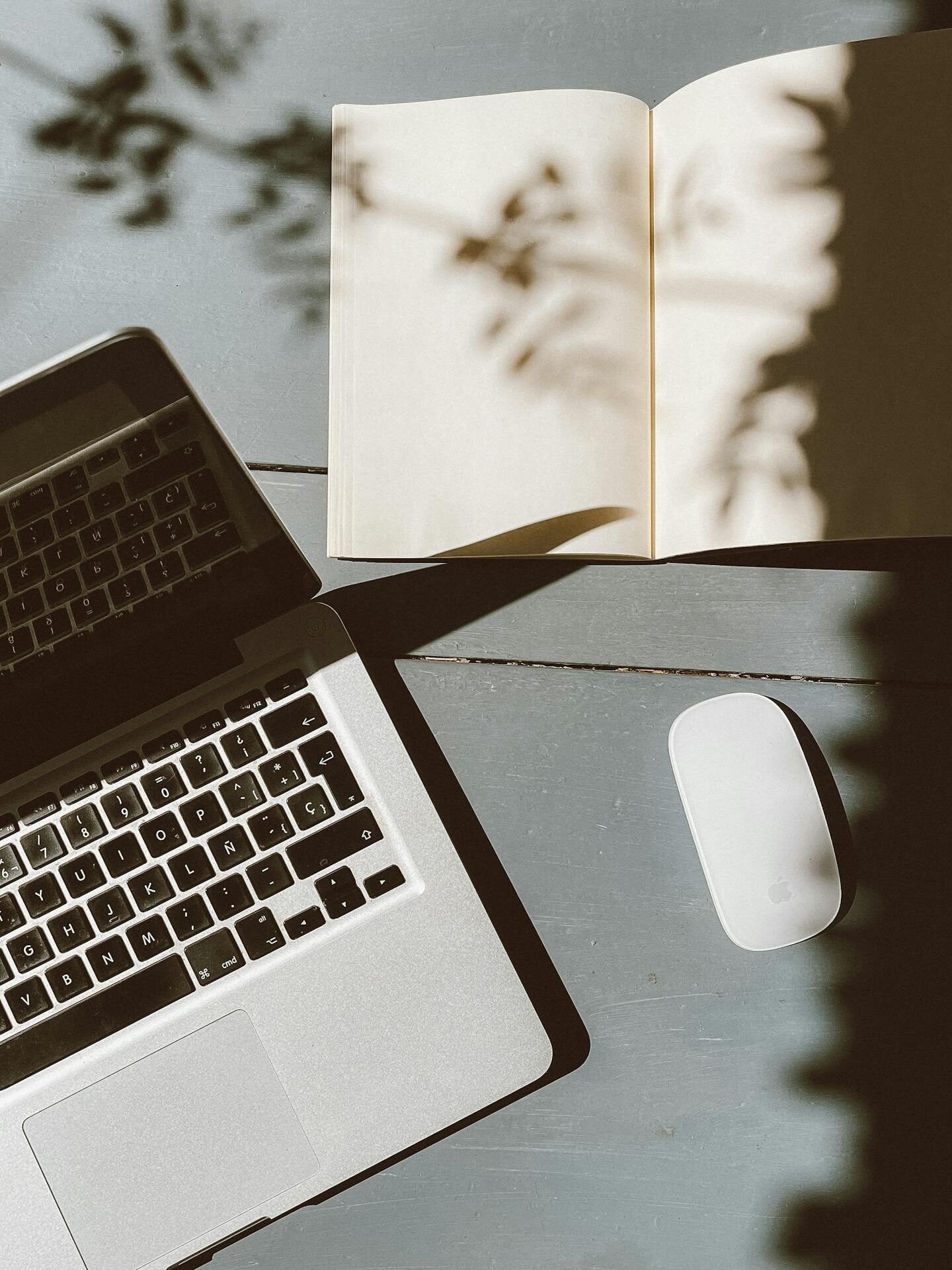 A sunlit workspace featuring a laptop, open notebook, and wireless mouse casting artistic shadows.